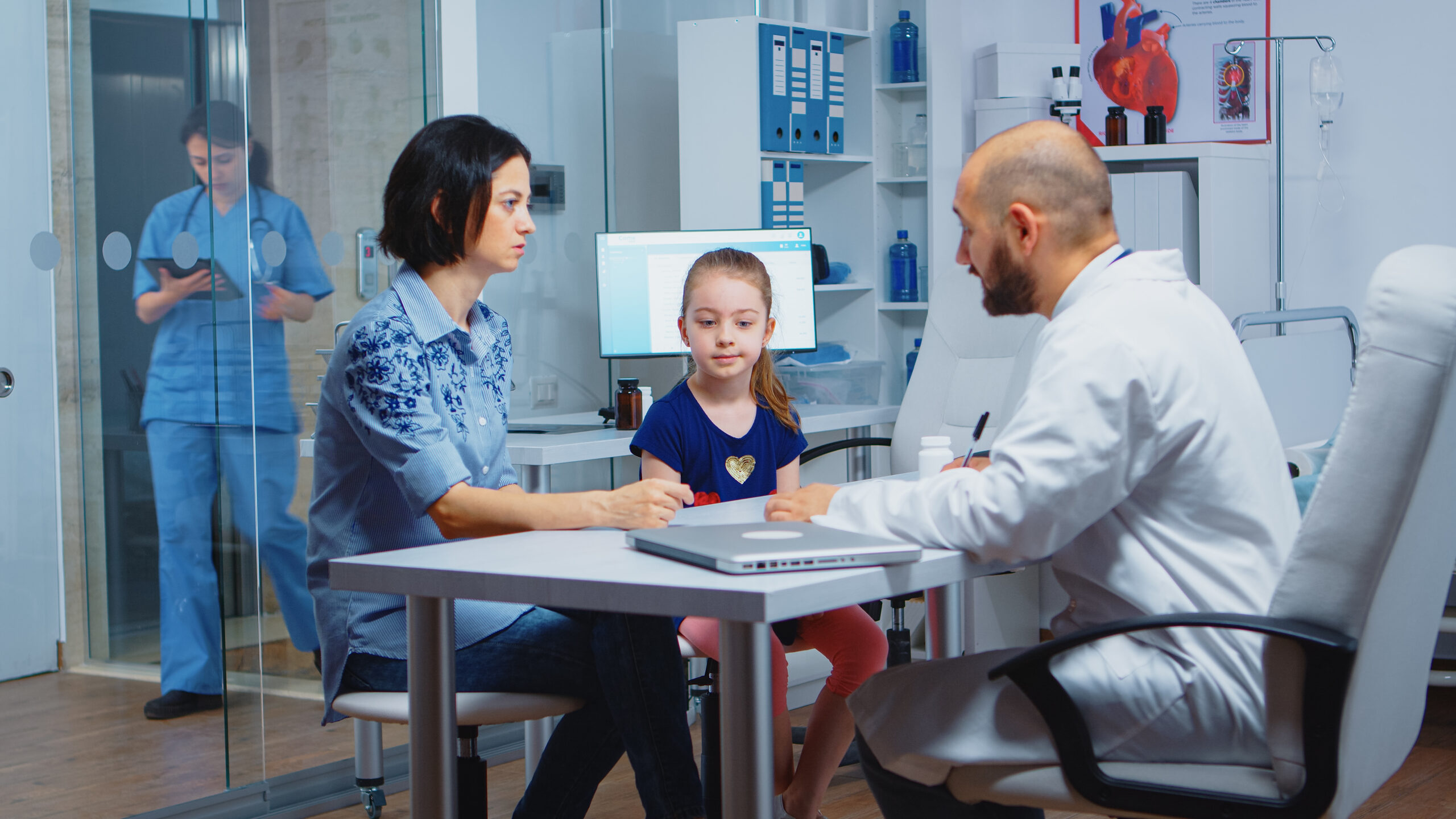 Doctor examining a patient in a clinic while the patient's young daughter sits beside them, representing family-focused primary care services.